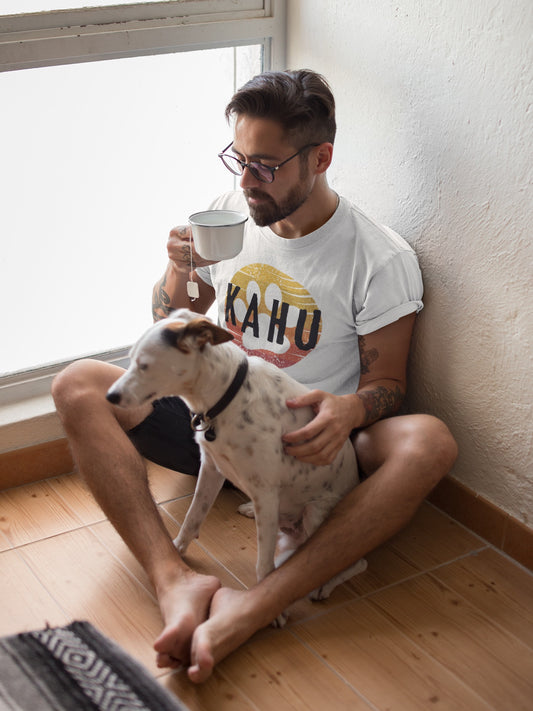 Man sitting on the floor holding a mug and a dog, wearing a white t-shirt with 'KAHU' text and sunset paw print graphic design - Island Drops t-shirt collection | Unisex t-shirts