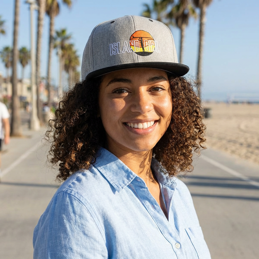 Woman wearing a gray and black cap with 'ISLAND GIRL' text and a surfer girl and sunset graphic design - Island Drops hat collection | Unisex snapback cap